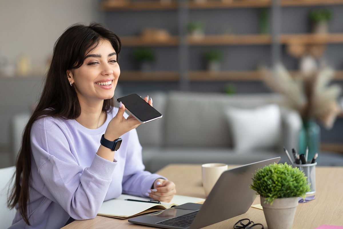 Digital Speaker Application. Portrait of young smiling woman sitting at table, talking on mobile phone, using laptop and virtual voice assistant for online search, to record reminder, dictate message